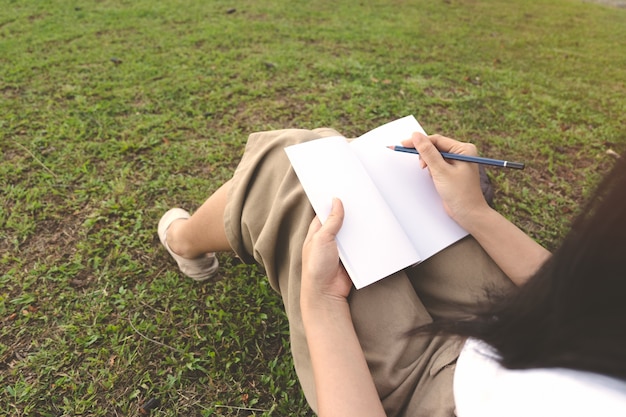 Young woman writing on notebook with pencil on grass in outdoor park.