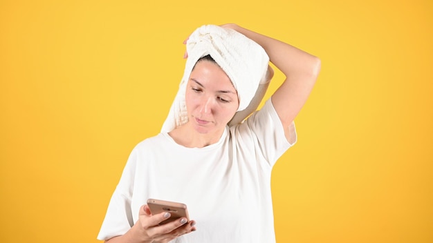 Young woman on yellow background with a towel on her head looks at the smartphone camera Woman holding cellphone