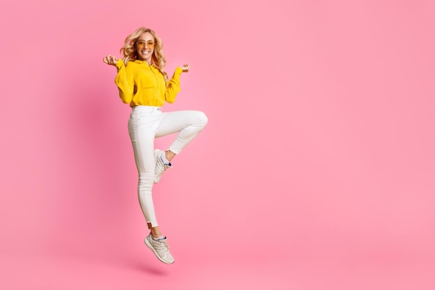 Photo a young woman in a yellow hoodie and white jeans is jumping in the air with a big smile on her face