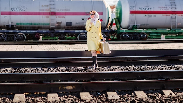 Young woman in yellow raincoat and dark glasses with suitcase in her hands crosses railroad tracks