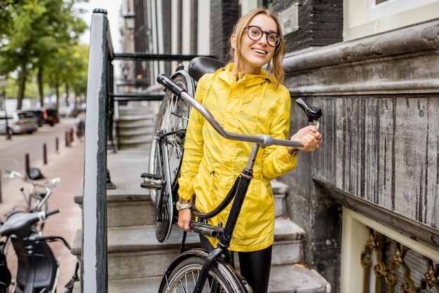 Young woman in yellow raincoat taking down a bicycle on the stairs near the beautiful residential house in Amsterdam