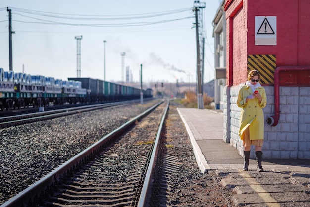 A young woman in yellow raincoat with phone in her hands on platform of railway station