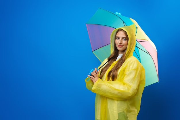 Young woman in yellow raincoat with rainbow umbrella against blue background in studio