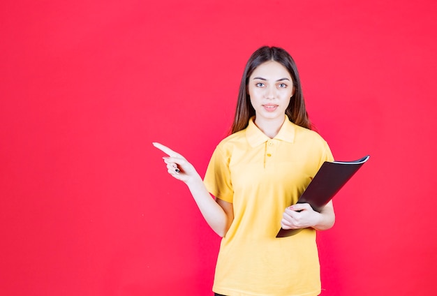 Young woman in yellow shirt holding a black folder, pointing and calling her colleague