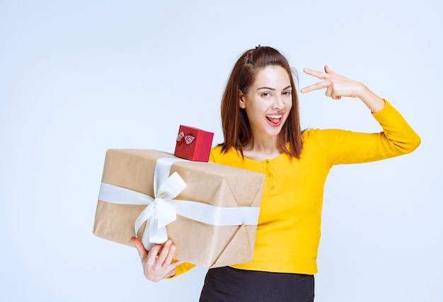 Young woman in yellow shirt holding a red and a cardboard gift boxes and showing positive hand sign