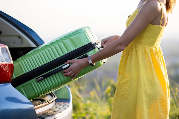 Young woman in yellow summer dress taking green suitcase from car trunk. Travel and vacations concept.