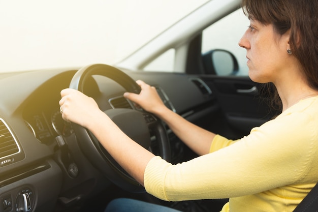 Young woman in yellow sweater and blue jeans holding the steering wheel of a car from inside the cabin