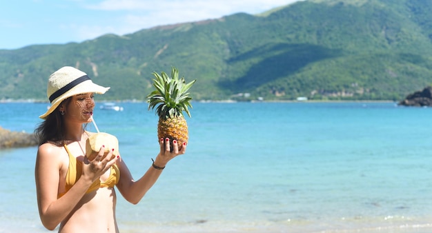 Young woman in yellow swimsuit drinking coconut milk and holding pineapple on sandy beach