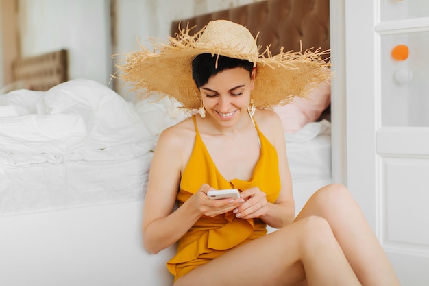 Young woman in yellow swimsuit and straw hat using smartphone sits by a bed in a hotel room