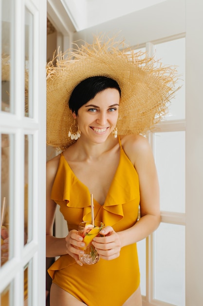 Young woman in yellow swimsuit and straw hat with a cocktail smiling in a window