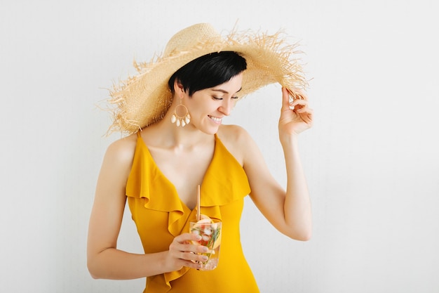 Young woman in yellow swimsuit and straw hat with a cocktail on white background