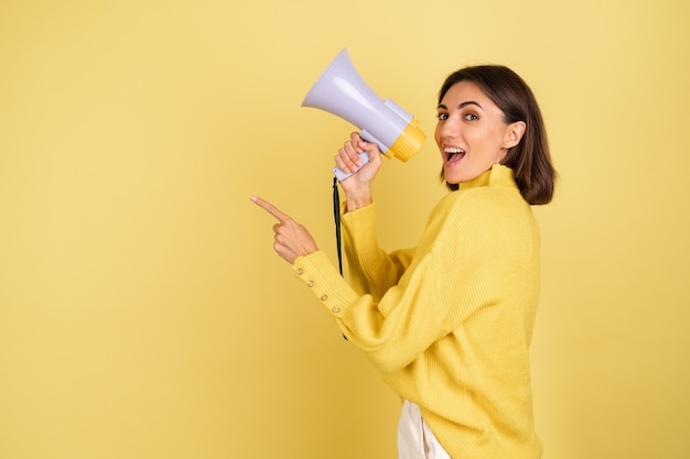 Young woman in yellow warm sweater with megaphone speaker screaming to the left pointing index finger