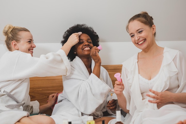 The young women in bathrobes are sitting on the bed and using skincare products