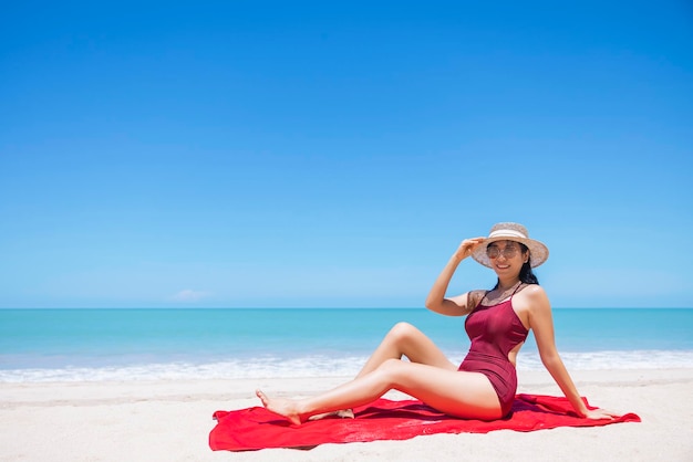 Young women in bikini and straw hat sit on tropical beach enjoying looking view of beach ocean