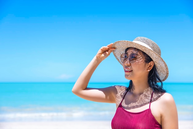 Young women in bikini and straw hat stand on tropical beach enjoying looking view of beach ocean