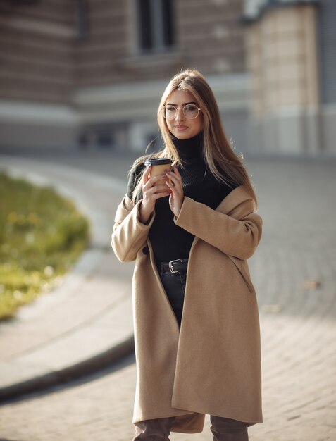 Young women dressed in a stylish coat drinks coffee on a background of urban architecture