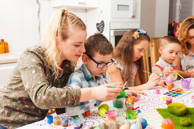 Young women heping kids creating spring decor for Easter. Mother and her son holding brushes painting colorful egg