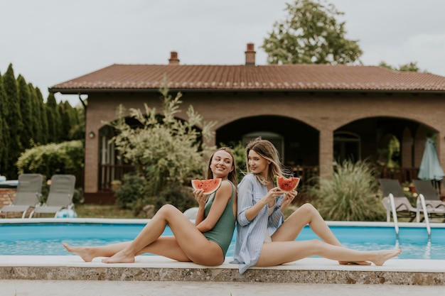 Young women sitting on by the swimming pool and eating watermelon in the house backyard