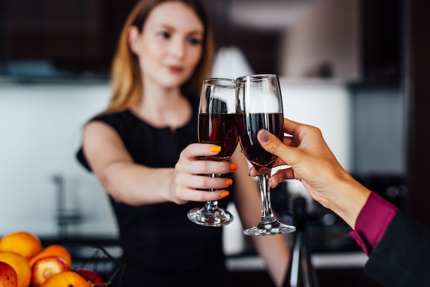 Young women wearing elegant black dress holding a bottle of red wine and a glass standing at kitchen bar looking at her female friend.