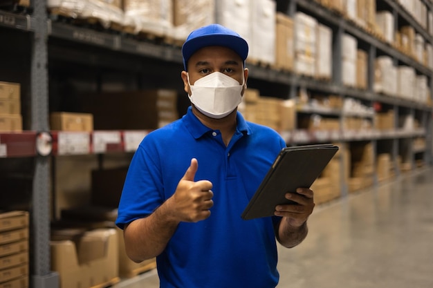 Young worker in blue uniform wearing protection mask checklist manage parcel box product in warehouse. Asian man employee using tablet working at store industry. Logistic import export concept.