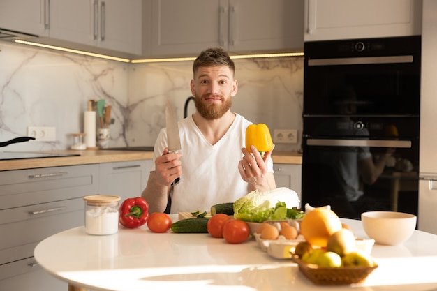 Young worried man sit at table and hold pepper and knife. Table with vegetables, fruits, eggs and salt. European bearded guy. Interior of kitchen in modern apartment. Concept of healthy nutrition