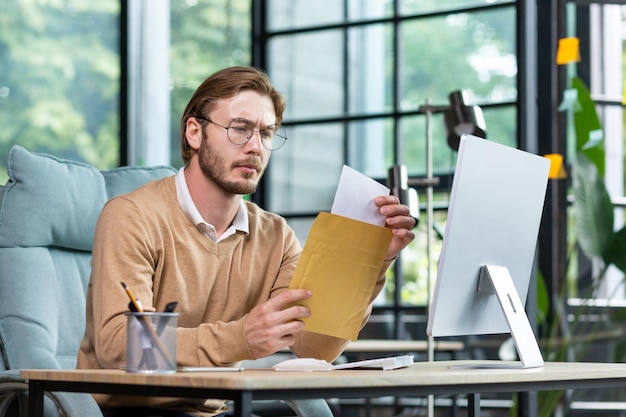 A young worried man sits at the desk in the office and opens an envelope with a letter papers