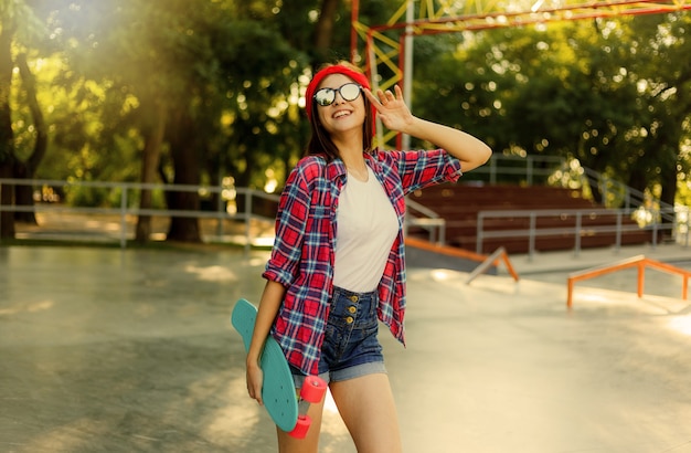 Youth sport. Young attractive hipster woman wearing a red plaid shirt and denim shorts holding a skateboard in a skatepark. Summertime