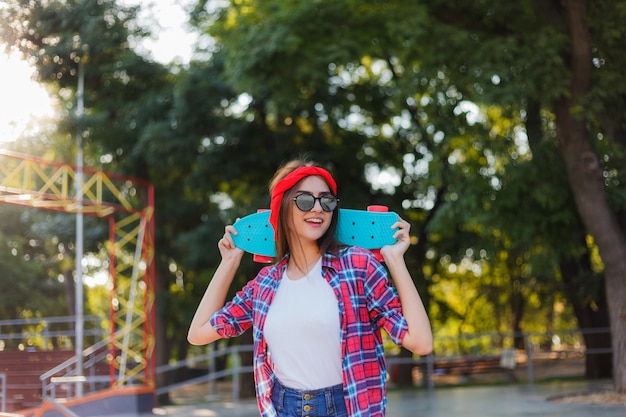 Youth sport. Young hipster woman wearing a red plaid shirt and denim shorts holding a skateboard in a skatepark. Summertime