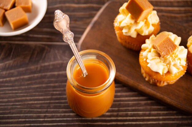 Yummy salted caramel with spoon from glass jar at wooden table and cupcakes on the background