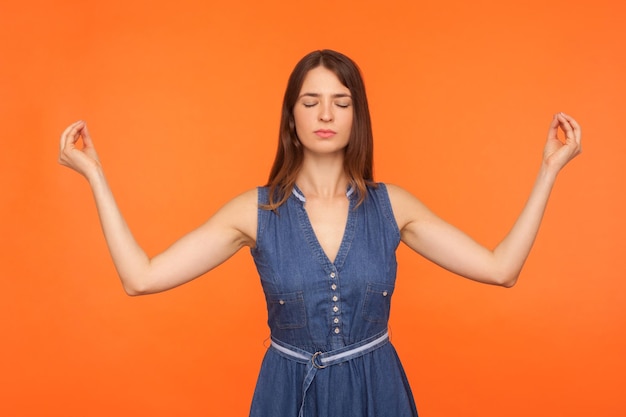 Zen practice breath technique Brunette woman in denim dress doing yoga exercise holding hands in mudra gesture and meditating feeling tranquil relaxed studio shot isolated on orange background