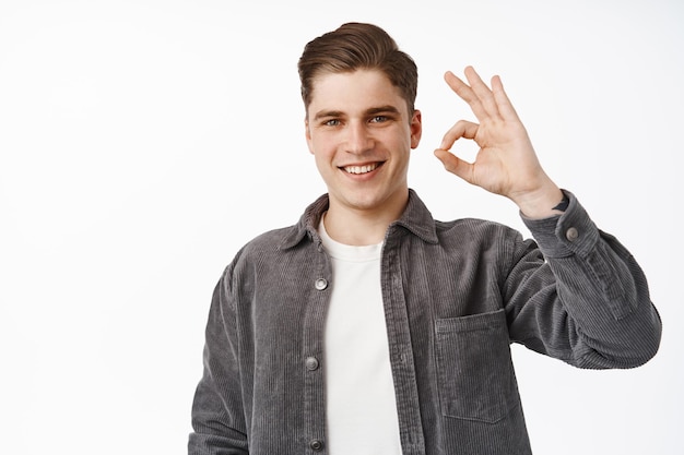 Zero no problems. Smiling young man shows okay OK sign, looks confident, assured all under control, no problem, satisfied with result, approve and like, praise you, white background.