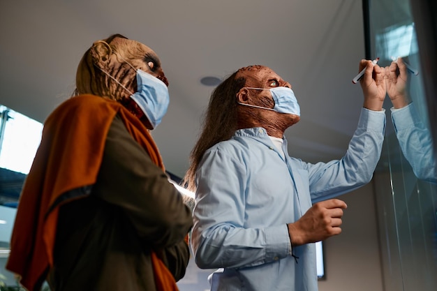 Zombie businessmen in face masks brainstorming discussing project, searching strategy drawing on glass board. Work in corporate office during pandemic concept