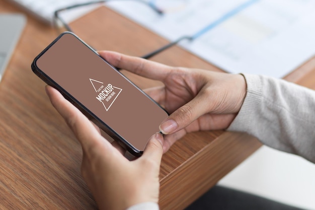 Mock up blank mobile screen by holding with 2 hands on the wooden desk with office supplies