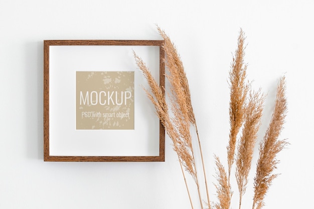 Square picture frame mockup on white wall with dry plant decorations