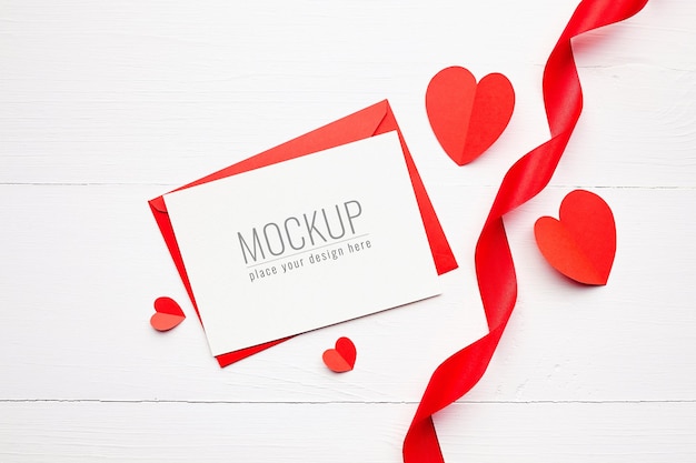 Valentines day card mockup with gift box, red ribbon and paper hearts on white wooden surface