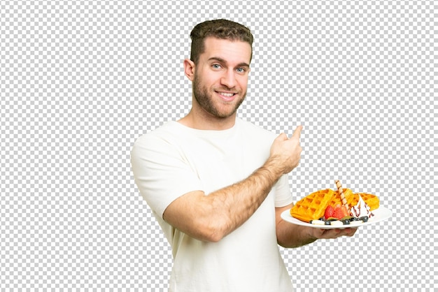 Young handsome blonde man holding waffles over isolated chroma key background pointing back