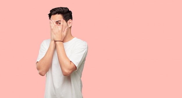 young handsome man with a scared, frightened expression, covering eyes with both hands, peeking