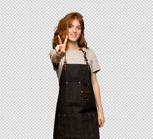 Young redhead woman with apron smiling and showing victory sign