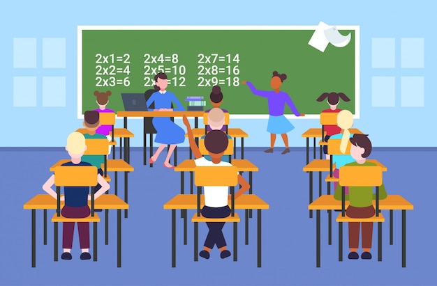 female teacher with pupils sitting at desks looking at schoolgirl solving math problem at chalkboard school lesson education concept modern school classroom interior  full length horizontal