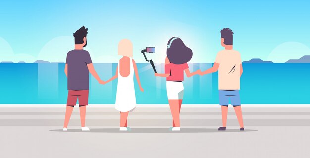 people group on beach using selfie stick taking photo on smartphone camera summer vacation concept rear view friends standing together seaside sea holiday ocean travel full length horizontal