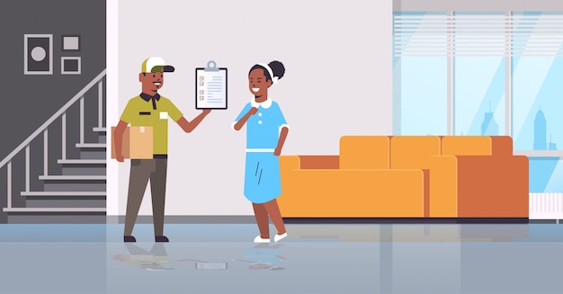 postman in uniform holding cardboard parcel box   courier showing clipboard to woman recipient express delivery service concept modern living room interior  full length