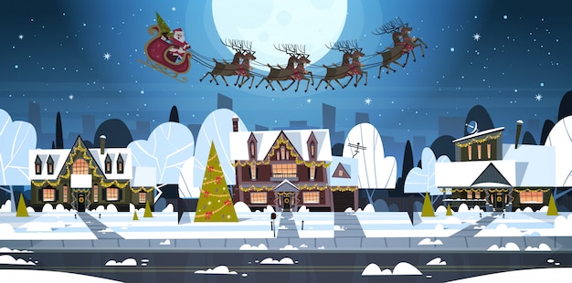 Santa Flying In Sledge With Reindeers In Sky Over Village Houses, Merry Christmas And Happy New Year Banner Winter Holidays Concept